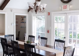 A bright dining room with a long wooden table surrounded by black chairs. The table is decorated with a beige runner, candles, and a small plant centerpiece. There is a chandelier hanging above the table, a window with blinds, and a glass door with an exit sign above it.