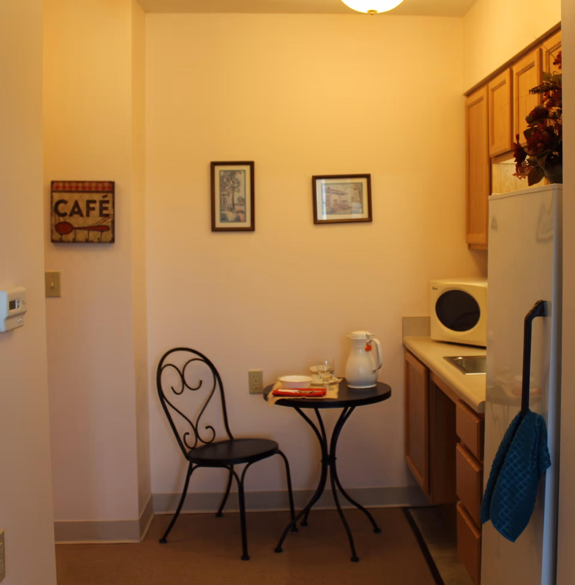 A small kitchen area with wooden cabinets, a white microwave, a white refrigerator with a blue towel hanging on the handle, and a small round black table with a matching black chair. On the table, there is a white pitcher, a glass, a bowl, and a red napkin. The walls are light-colored with two framed pictures and a decorative sign that says 'CAFÉ'.