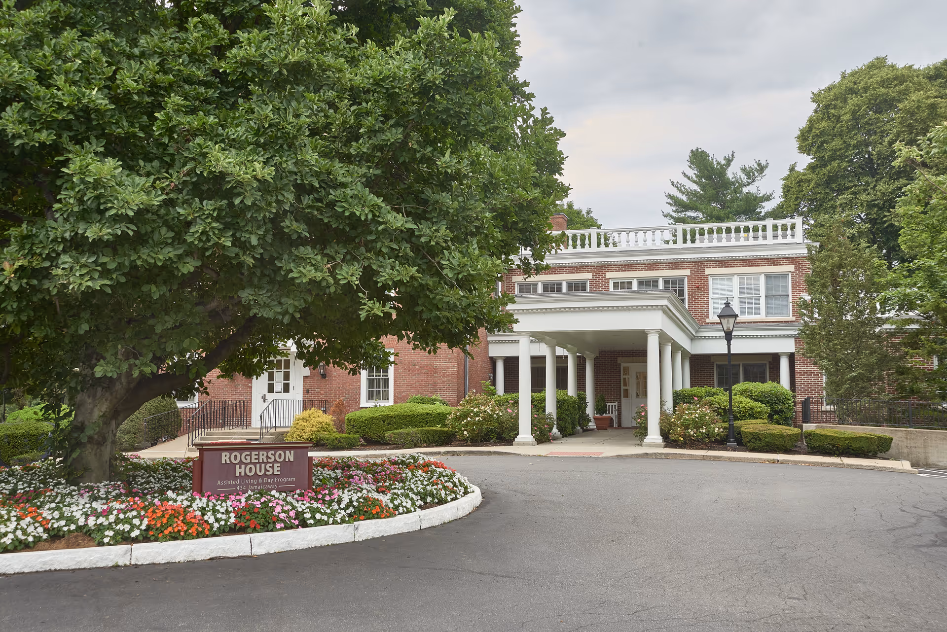 Front exterior view of Rogerson House Memory Care Assisted Living & Day Program building with a large tree and flower bed in front, a covered entrance with white columns, and well-maintained shrubs and greenery around the building.