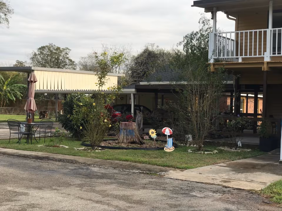 Outdoor area of a senior living facility with a small garden featuring a tree stump, decorative flower and mushroom stakes, and surrounding plants. There is a patio table with chairs and a closed umbrella on the left side. A covered parking area with cars is visible in the background, and part of a two-story building with a balcony is on the right.