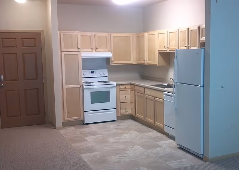 A clean kitchen area in a senior living facility featuring light wood cabinets, a white electric stove with oven, a white refrigerator, a dishwasher, and a double sink. The floor is tiled and there is a wooden door to the left.