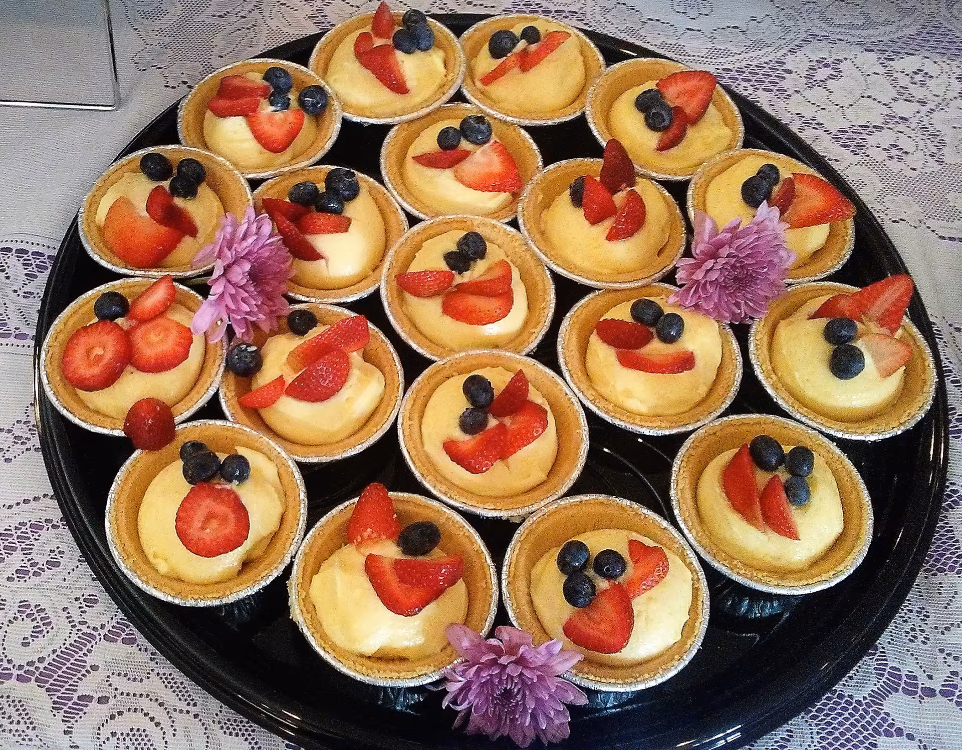 A black tray holding multiple small dessert cups filled with creamy yellow custard, each topped with fresh blueberries and sliced strawberries, decorated with purple flowers, placed on a lace tablecloth.