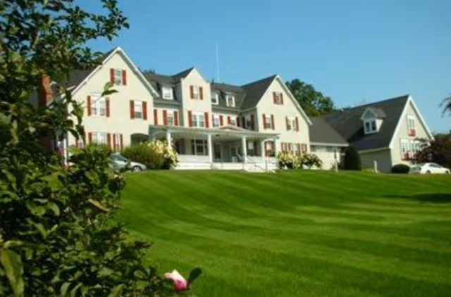 Large white multi-story residential building with a wide porch set behind a manicured green lawn under a clear blue sky.