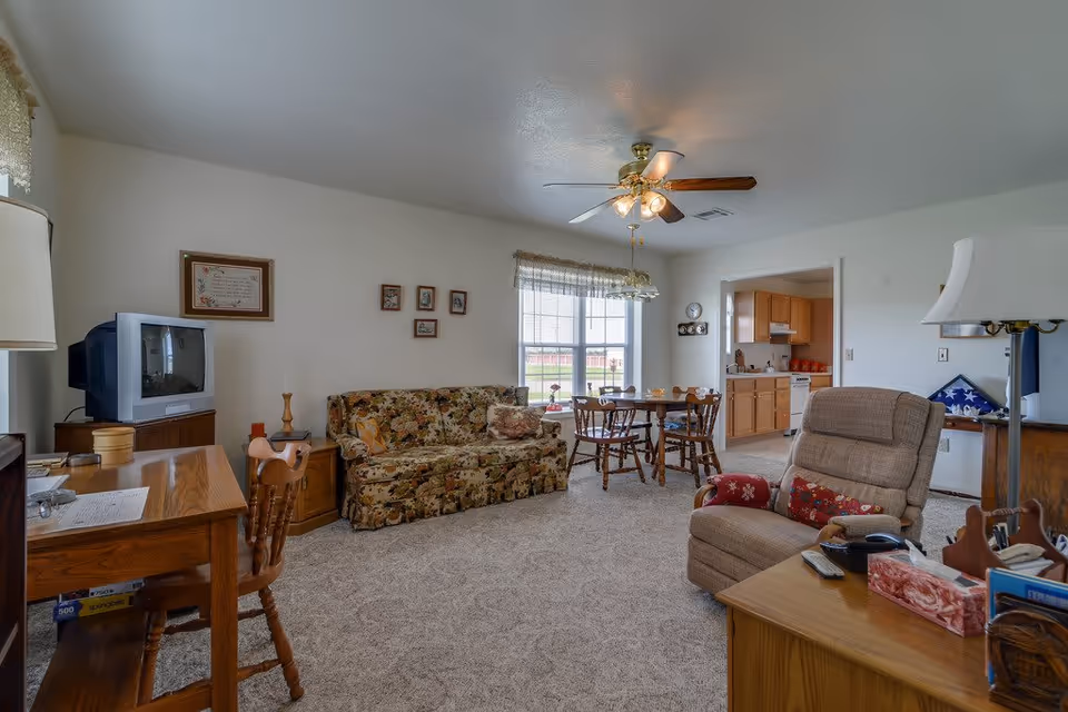A cozy senior living room with a floral patterned sofa, a wooden dining table with four chairs near a window, a beige recliner chair, a wooden desk with a chair, and a TV on a stand. The room has beige carpet, white walls, and a ceiling fan with lights. A kitchen area is visible through an open doorway.