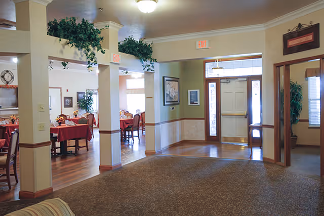 Interior view of a senior living facility showing a carpeted common area leading to a dining room with tables covered in red tablecloths and chairs. The space is decorated with green plants above the pillars and framed artwork on the walls. There is a door with glass panels and exit signs above the doorways.