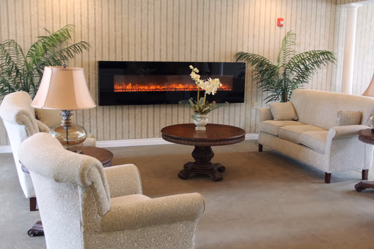 A cozy sitting area with upholstered chairs and a sofa facing a wall-mounted electric fireplace, a round wooden coffee table, lamps and potted plants.