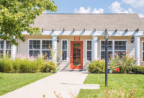 Front exterior of a single-story building with a red exit door, walkway, pergola, and landscaped garden.