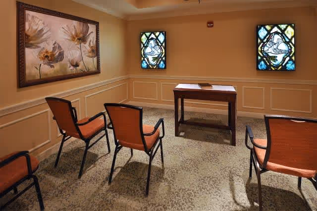 A small room with four chairs arranged in two rows facing a wooden table. The walls are beige with white panel molding, decorated with a large floral painting on the left wall and two stained glass windows on the front wall. The floor is carpeted with a patterned design.