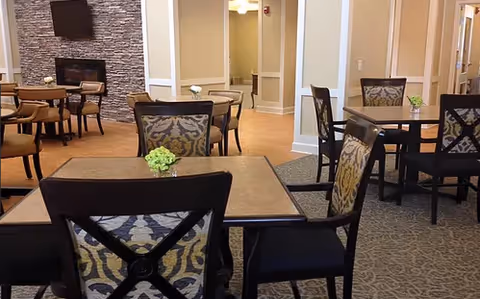 Communal dining area with multiple tables and patterned chairs, small green centerpieces, and a stone fireplace with a wall-mounted TV.