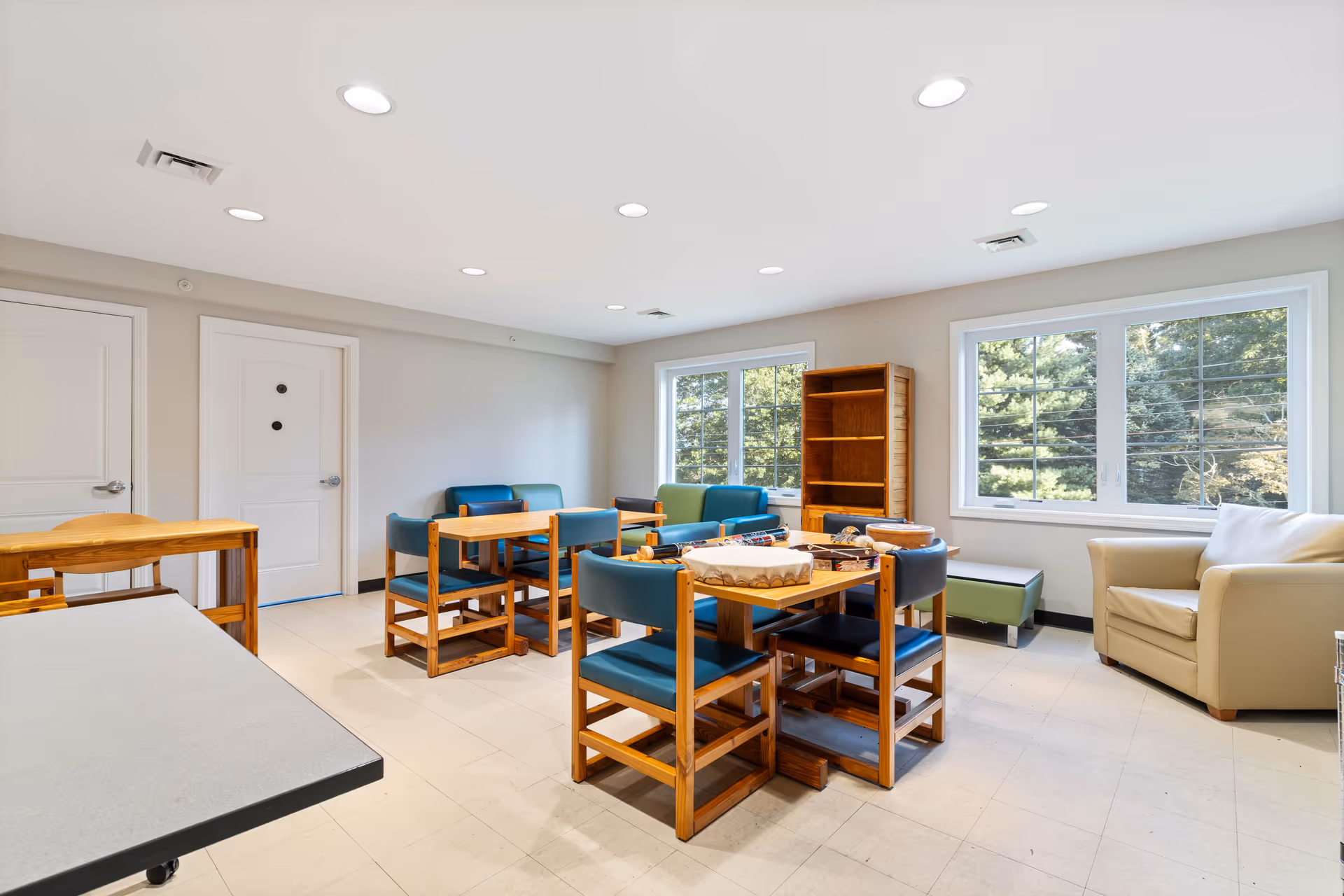 A bright room with multiple wooden tables and chairs with blue cushions, a beige armchair, a wooden bookshelf, and large windows showing green trees outside. The room has a tiled floor and recessed ceiling lights.