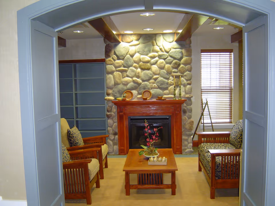 Cozy living room with a stone fireplace, wooden mantle, armchairs and a coffee table seen through an arched doorway.