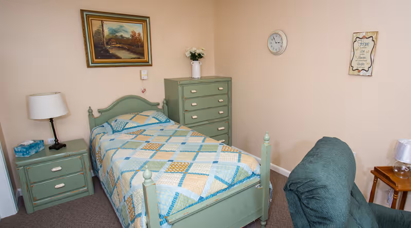 A small bedroom with a single bed featuring a patchwork quilt in shades of blue, green, and beige. The bed has a green wooden frame. To the left of the bed is a green nightstand with a lamp and a box of tissues. Behind the bed is a green chest of drawers with a white vase of flowers on top. On the wall above the bed is a framed landscape painting. A clock and a decorative wall plaque are hung on the beige walls. In the foreground, there is a green upholstered chair and a small wooden side table with a lamp.