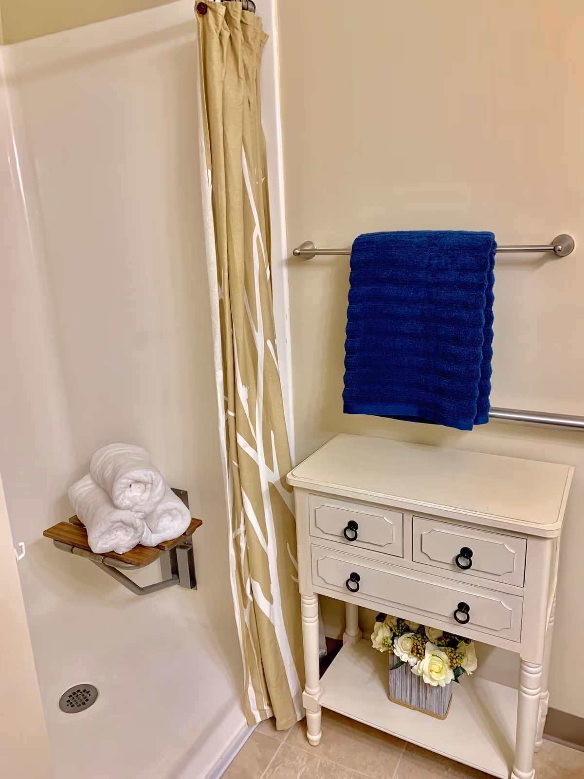 A bathroom shower area with a beige and white patterned shower curtain, a small wooden fold-down seat holding three rolled white towels, a white cabinet with three drawers and a shelf underneath holding a flower arrangement, and a silver towel rack mounted on the wall with a folded dark blue towel.