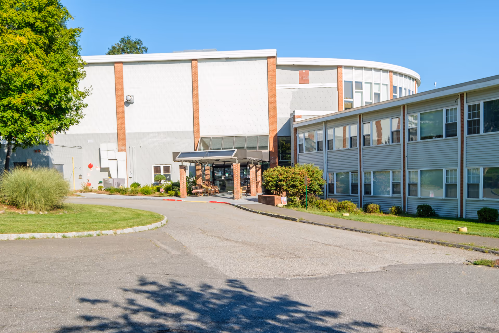 Exterior view of Tappan Zee Manor building with a driveway leading to the entrance. The building has a combination of brick and siding with multiple windows, surrounded by green grass, bushes, and a tree on the left side under a clear blue sky.