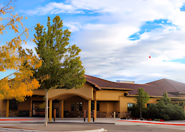 Front exterior of a single-story assisted living building with a covered entrance, trees, and landscaping under a partly cloudy sky.