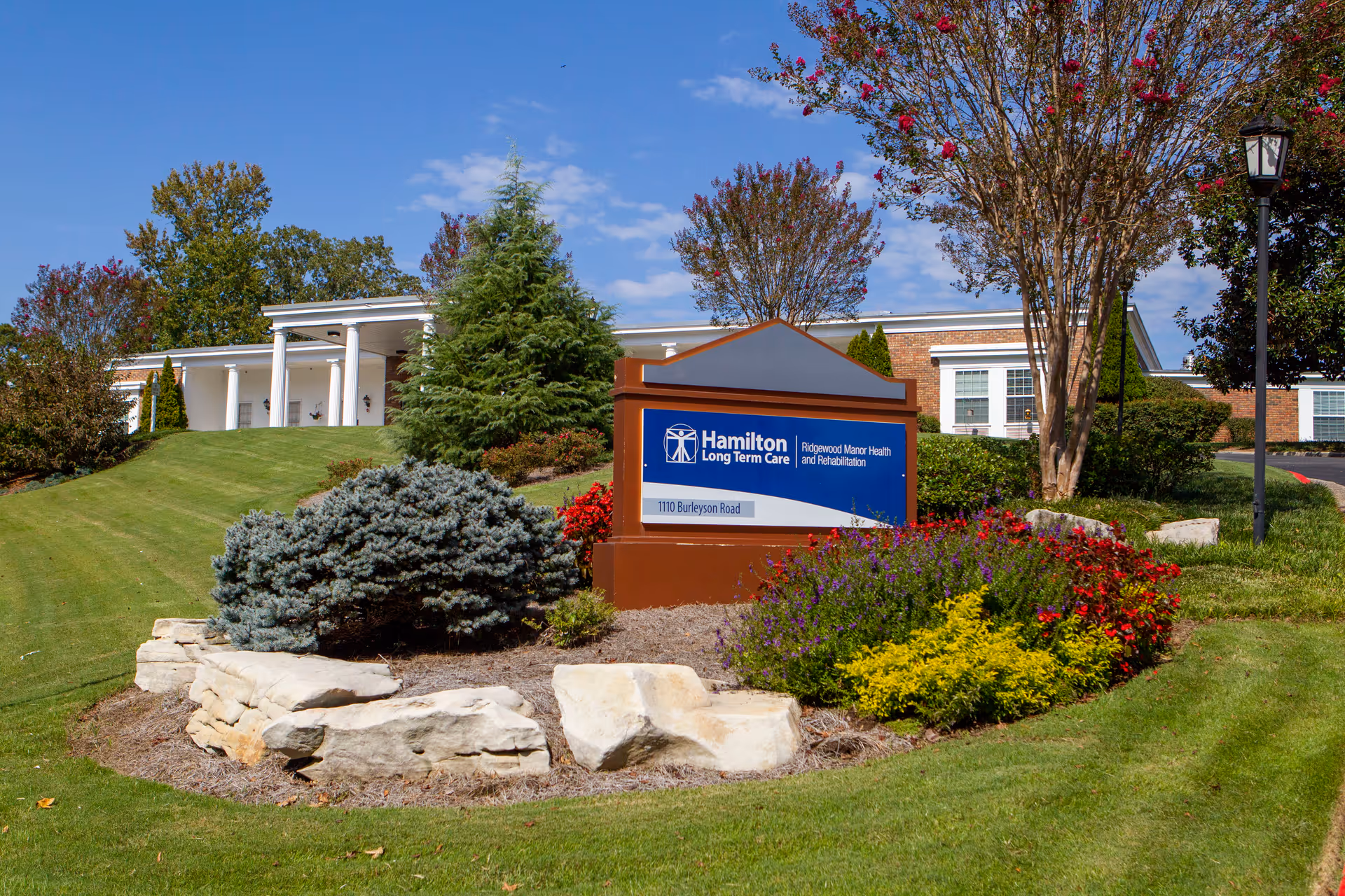A landscaped front lawn with a Hamilton Long Term Care sign for Ridgewood Manor Health and Rehabilitation and the facility building in the background.