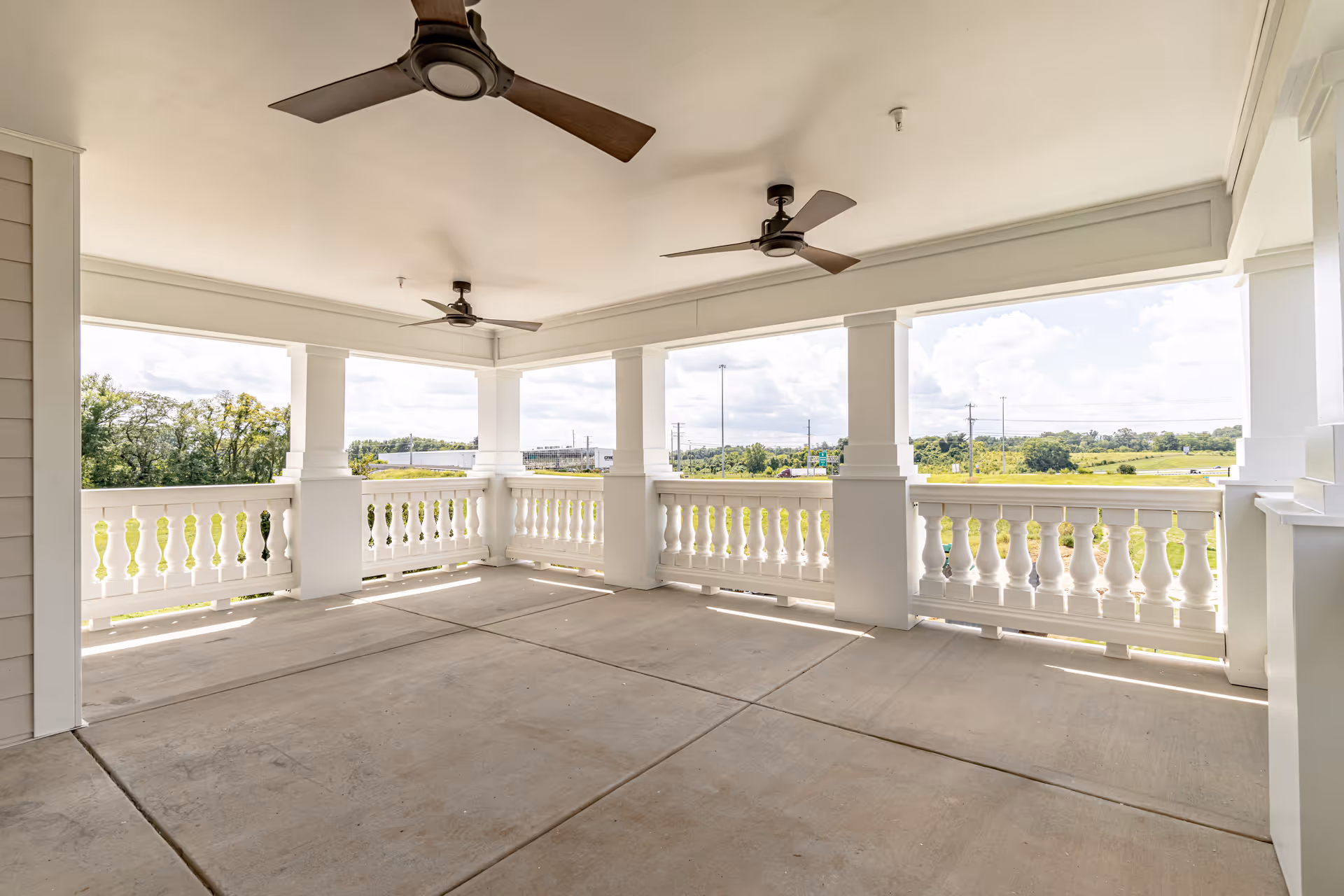 Covered outdoor patio area with white railings and columns, ceiling fans, and a view of green fields and trees in the background under a partly cloudy sky.