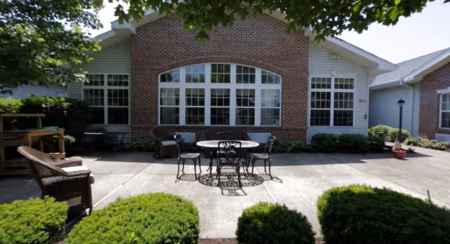 Outdoor patio area with a round metal table and four chairs on a concrete surface, surrounded by green bushes. The background shows a brick building with large windows and a white trim, along with some potted plants and outdoor furniture.