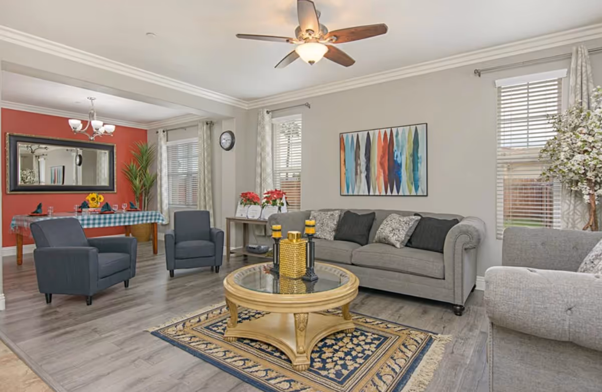 A bright and cozy living room area with a gray sofa, two gray armchairs, a round wooden coffee table with decorative items, and a patterned rug. The room has light gray walls with a colorful abstract painting, large windows with blinds and curtains, and a ceiling fan with a light. In the background, there is a dining area with a table covered with a checkered tablecloth, chairs, a large mirror on a red accent wall, and a potted plant.