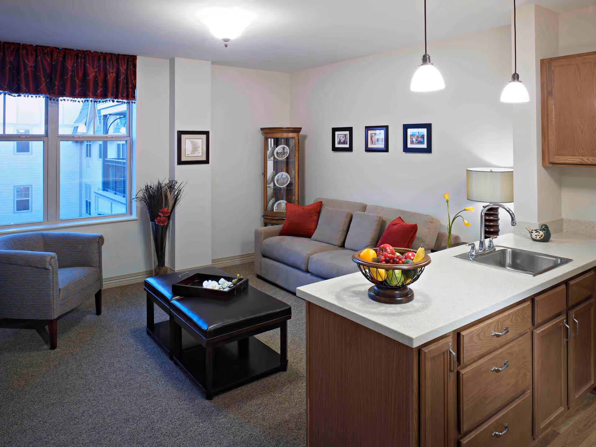 A cozy living room area with a gray sofa adorned with red and gray cushions, a wooden coffee table with a black tray, and a single armchair near a window with red curtains. Adjacent to the living room is a kitchen counter with a sink, a lamp, and a fruit basket. The walls are decorated with framed pictures and a glass display cabinet with plates.