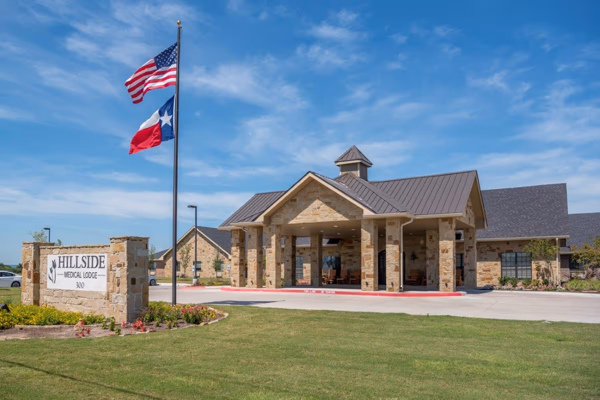 Exterior view of Hillside Medical Lodge building with stone facade and a covered entrance. Two flagpoles display the American and Texas flags. The building is surrounded by a well-maintained lawn and landscaping under a blue sky with some clouds.