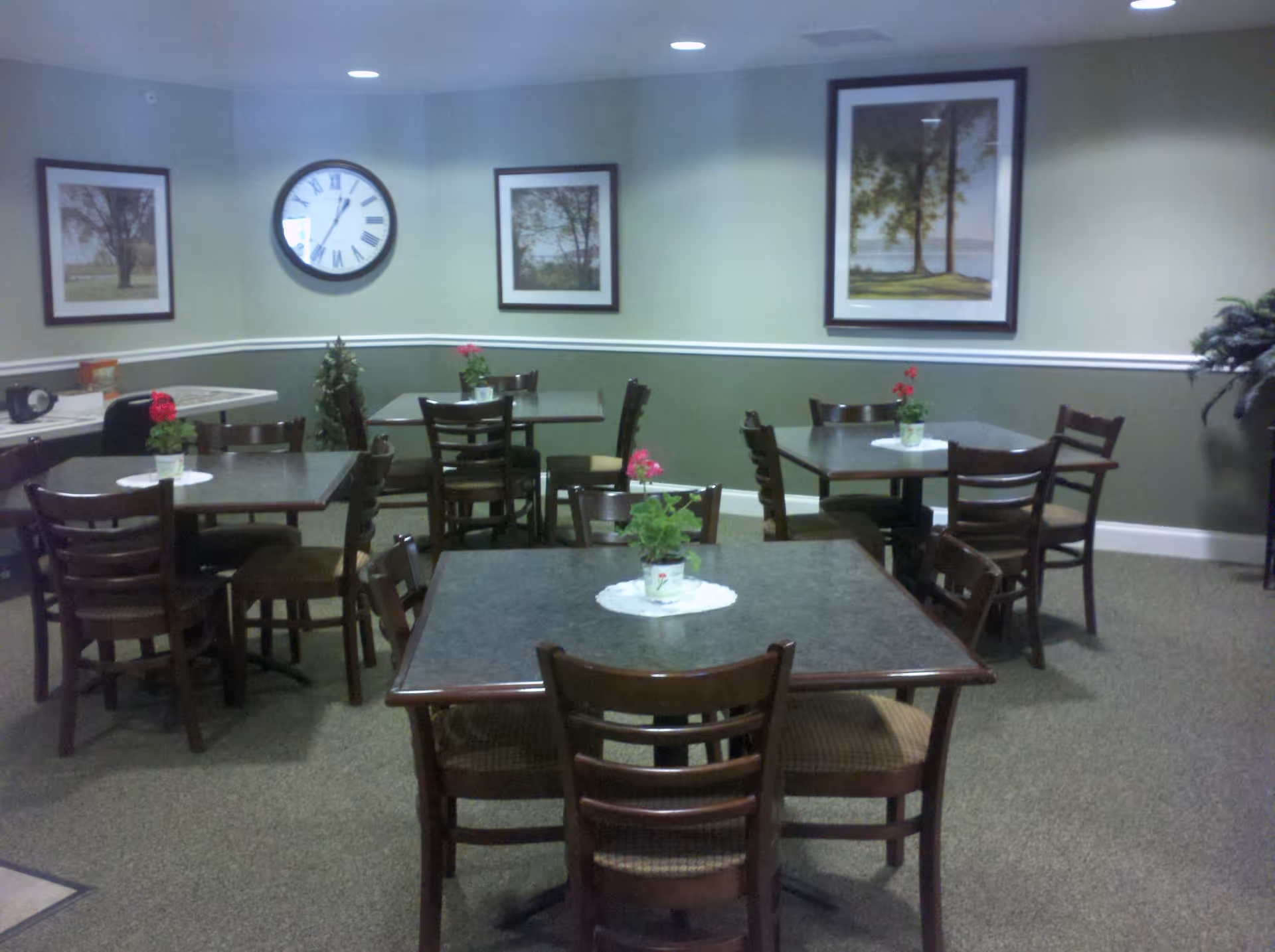 Communal dining room with several square tables and wooden chairs, small potted flowers on doilies on the tables, and a wall clock and framed landscape prints.
