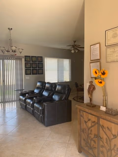 Interior view of a living room with three black leather recliner chairs arranged in a row facing away from a sliding glass door with vertical blinds. The room has tiled flooring and neutral-colored walls. On the right side, there is a wooden cabinet with decorative carvings, a vase with three sunflowers, a small statue, and framed certificates hanging on the wall above it. A ceiling fan with lights is visible in the background.