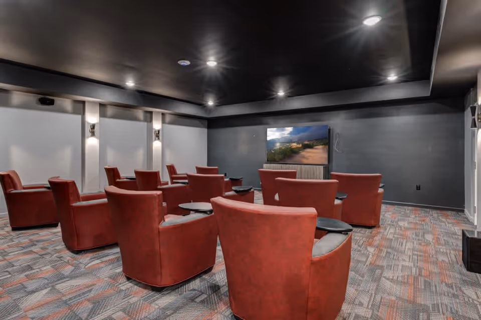 A modern media room with multiple red armchairs arranged in rows facing a large flat-screen TV mounted on a dark wall. The room has a dark ceiling with recessed lighting and patterned carpet flooring.
