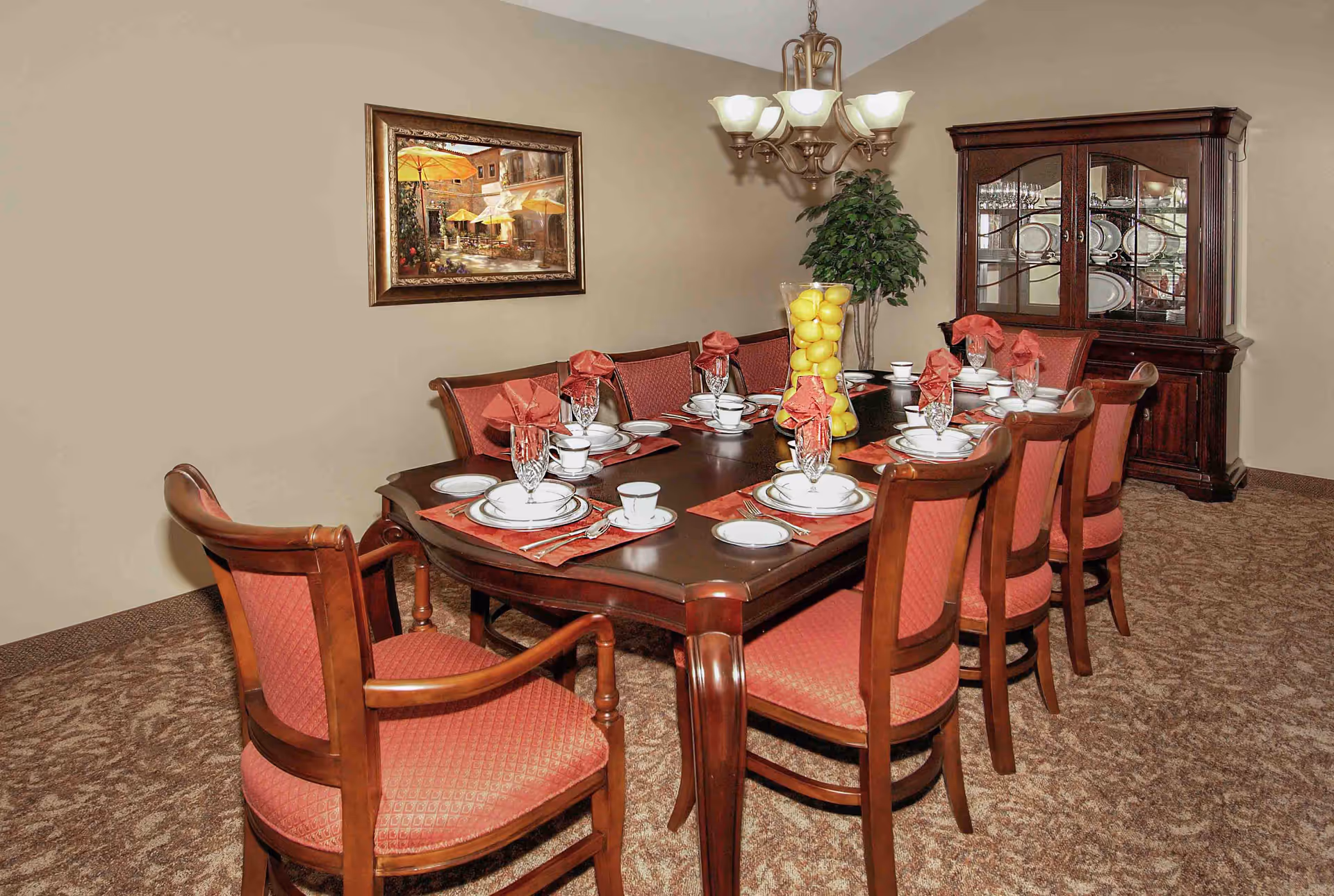 A formal dining room with a dark wooden table set for eight people. Each place setting includes white plates, cups, crystal glasses, and red napkins folded in a decorative manner. The chairs have wooden frames with red upholstered seats and backs. A large glass vase filled with yellow lemons serves as a centerpiece. In the background, there is a wooden china cabinet displaying plates and glassware, a green potted plant, a chandelier with five lights, and a framed painting on the beige wall.