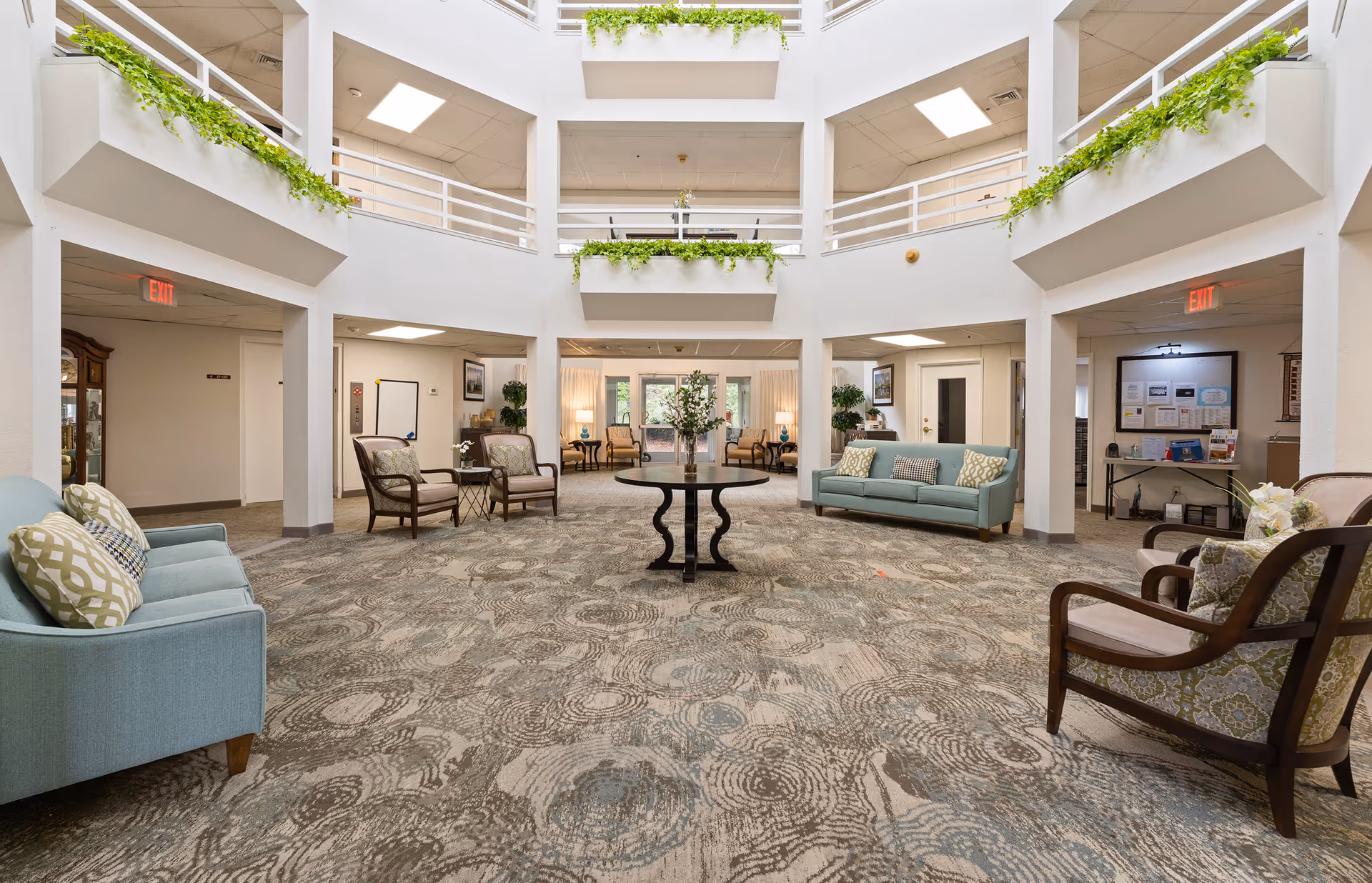 Spacious and well-lit common area in a senior living facility with patterned carpet, light blue sofas with decorative pillows, wooden armchairs with patterned cushions, a round table with a flower arrangement in the center, and greenery hanging from the upper balcony railings.