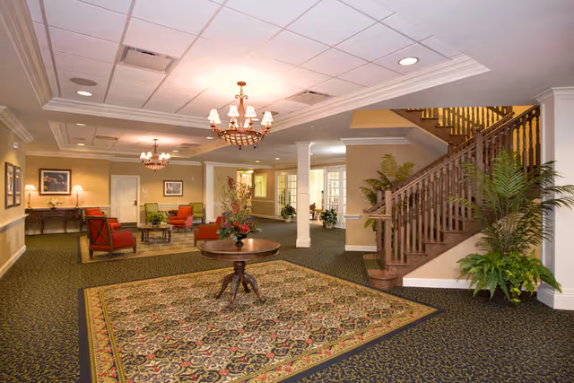 Spacious interior common area with patterned carpet, a round wooden table with a floral arrangement in the center, multiple seating areas with red and green upholstered chairs, chandeliers hanging from the ceiling, a wooden staircase with railing, and several potted plants.