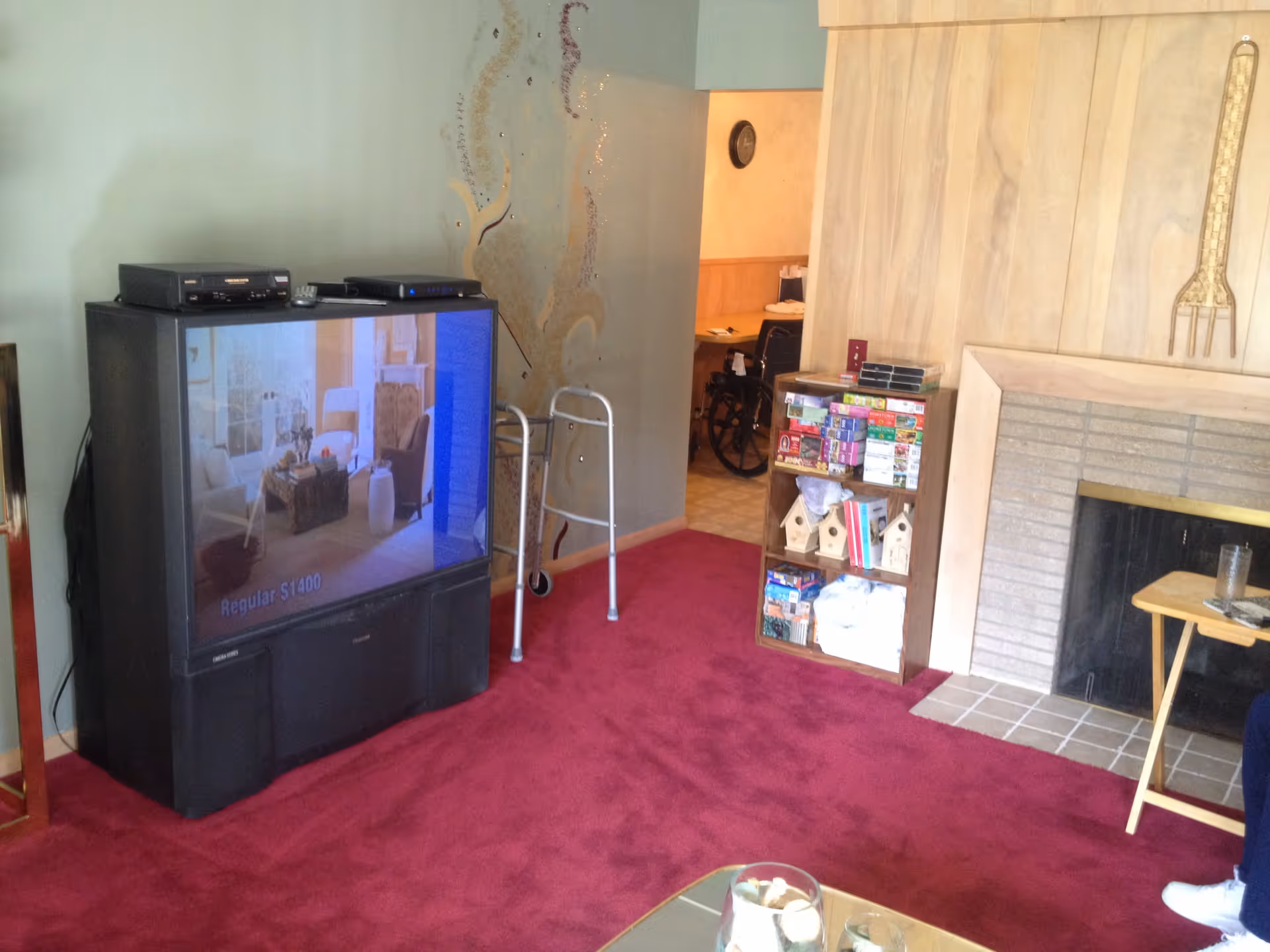A cozy living room with a large old-style television on the left, a walker positioned near the wall, a small wooden shelf filled with board games and birdhouses, and a fireplace with a light wood mantel on the right. The room has red carpet flooring and light-colored walls, with a small table holding a glass of water near the fireplace. A wheelchair is visible in an adjacent room through an open doorway.