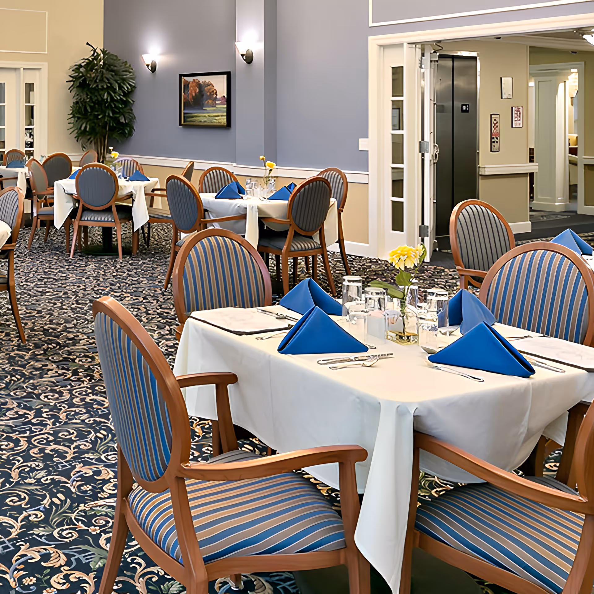 A dining room with multiple tables covered in white tablecloths, each set with blue folded napkins, glassware, and silverware. The chairs have wooden frames with blue and beige striped upholstery. The room has patterned carpet, light blue and beige walls, wall sconces, a framed landscape painting, and a potted plant in the corner. There is an open doorway leading to a hallway with an elevator.