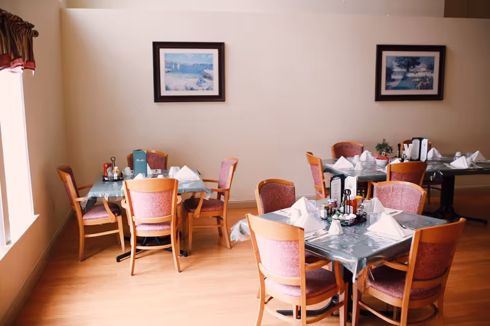 Dining room with several small tables set with napkins, condiments and wooden chairs beneath framed wall art and a window.
