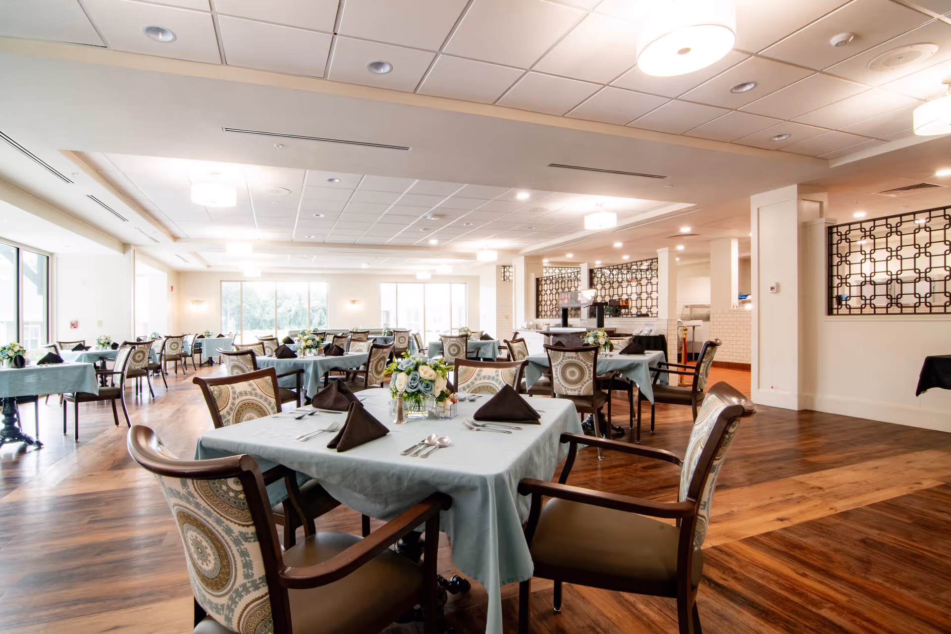 A spacious and well-lit dining room in a retirement community with multiple tables covered in light blue tablecloths, each set with silverware, napkins, and floral centerpieces. The room features wooden flooring, large windows allowing natural light, and decorative partitions.
