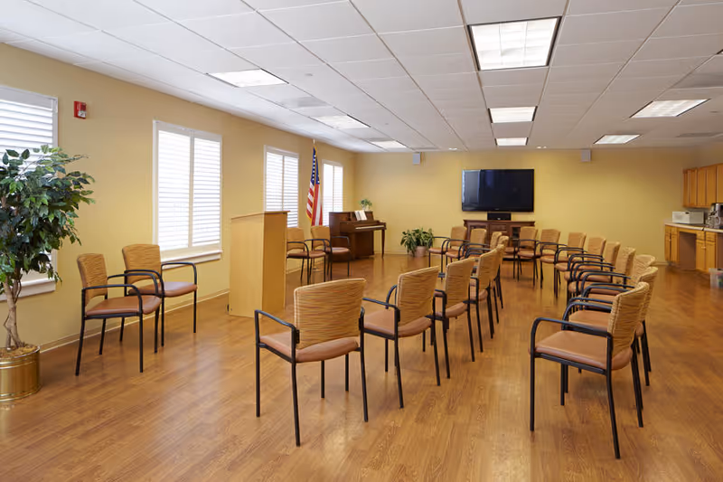 A spacious room with wooden flooring and beige walls, arranged with multiple chairs in rows facing a podium and a large flat-screen TV mounted on the wall. There is an American flag near the podium, a piano in the corner, several potted plants, and a kitchenette area with cabinets and appliances on the right side.