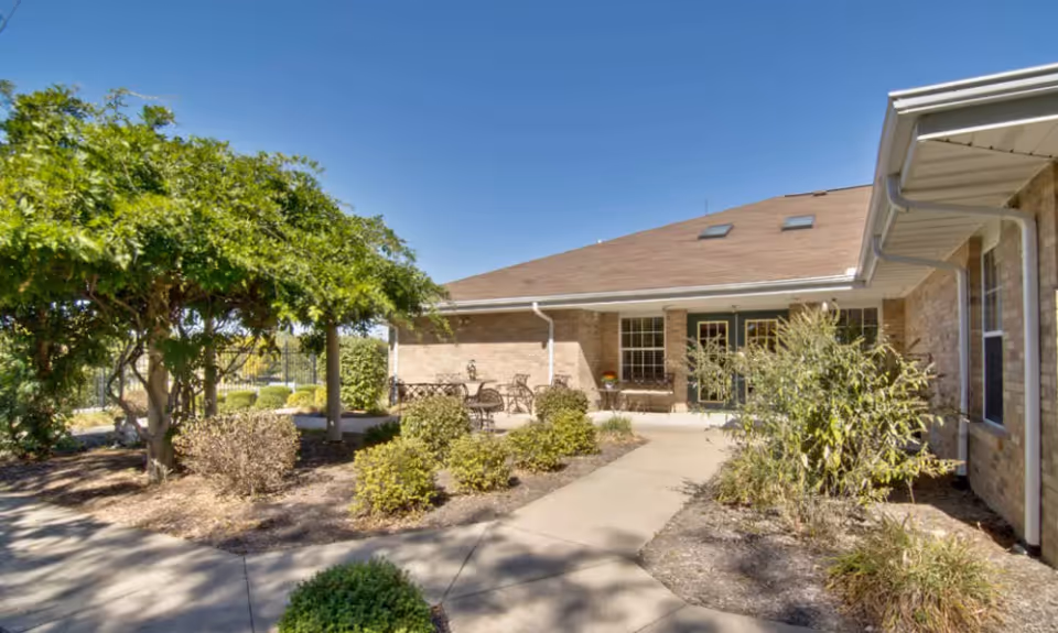 Outdoor courtyard and entrance of a single-story brick senior living building with a paved walkway, patio furniture, and landscaped shrubs under a clear blue sky.