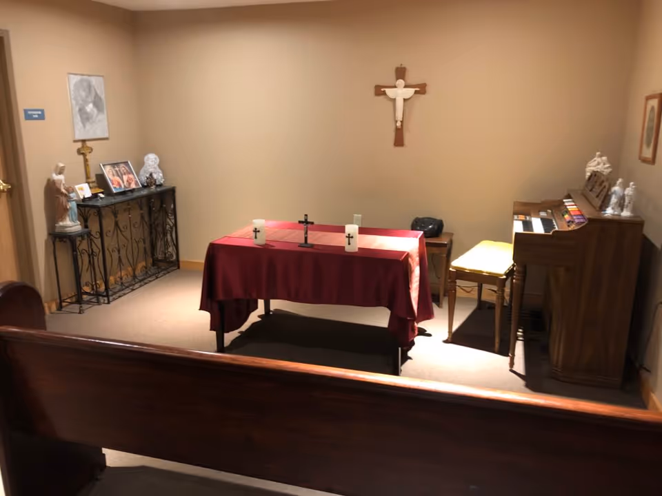 Small chapel room with a wooden pew in the foreground, a table covered with a red cloth holding two candles and a cross, a wooden organ with a yellow cushioned bench, religious statues and pictures on a side table, and a crucifix mounted on the beige wall.