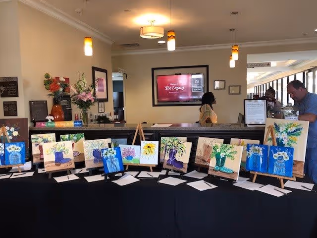 A display table covered with a black cloth showcasing multiple small paintings of flowers and plants on easels. Behind the table is a reception desk with two people interacting. A large screen on the wall behind the desk displays information about The Legacy at Crystal Falls. The room is well-lit with ceiling lights and decorated with flowers and framed pictures.