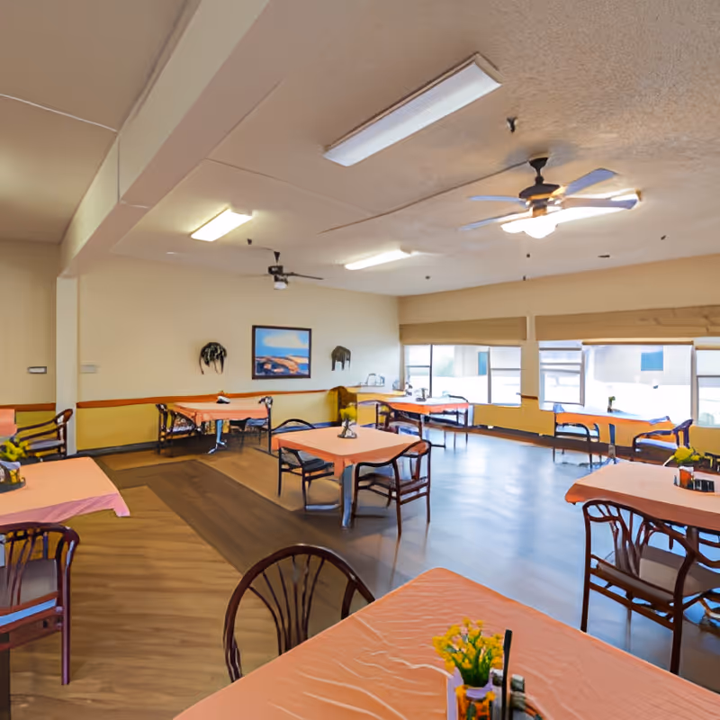 A spacious dining room with multiple tables covered in peach-colored tablecloths, each surrounded by wooden chairs. The room has large windows letting in natural light, ceiling fans, and framed artwork on the walls. Small flower arrangements are placed on each table.