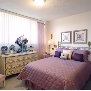 A cozy bedroom with a bed covered in a purple quilt and multiple pillows. There is a cream-colored dresser with decorative plates, a vase of flowers, and framed photos. The room has a window with vertical blinds and pink curtains, a bedside table with a lamp, and two framed floral pictures on the wall above the bed.