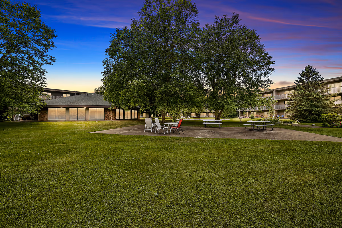 Courtyard of Heathers Estates with a large grassy lawn, patio tables and chairs beneath big trees and low-rise buildings at sunset.
