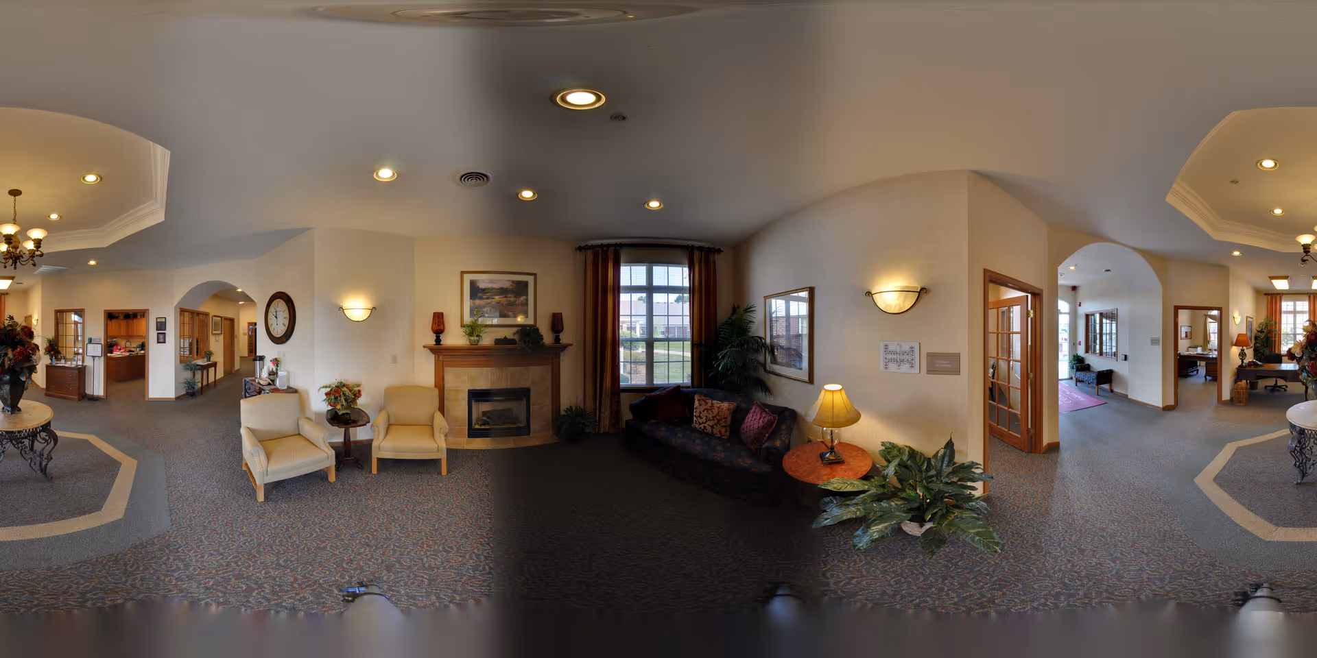 A spacious and warmly lit senior living facility common area featuring comfortable seating including armchairs and a sofa, a fireplace with decorative items on the mantel, large windows with curtains, indoor plants, and soft lighting from wall sconces and table lamps. The area has carpeted floors and leads into other rooms through arched doorways.