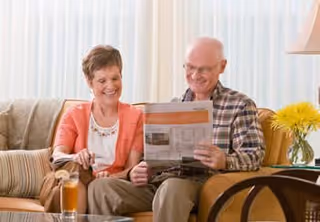 An elderly couple sitting on a beige couch in a living room, smiling and looking at a magazine together. There is a glass of iced tea on the table in front of them and a vase with yellow flowers on a side table nearby.