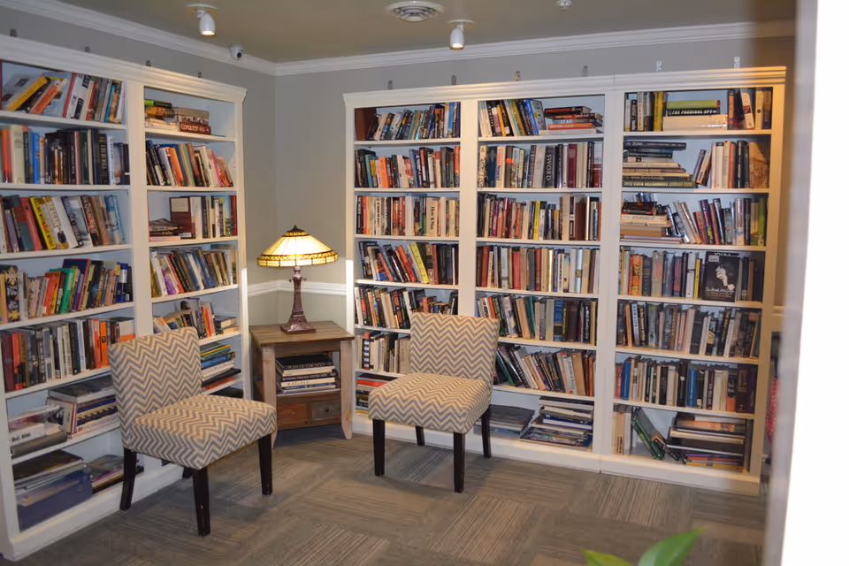 A cozy reading nook in a senior living facility featuring white built-in bookshelves filled with books, two patterned upholstered chairs, a small wooden side table with a decorative lamp, and soft overhead lighting.