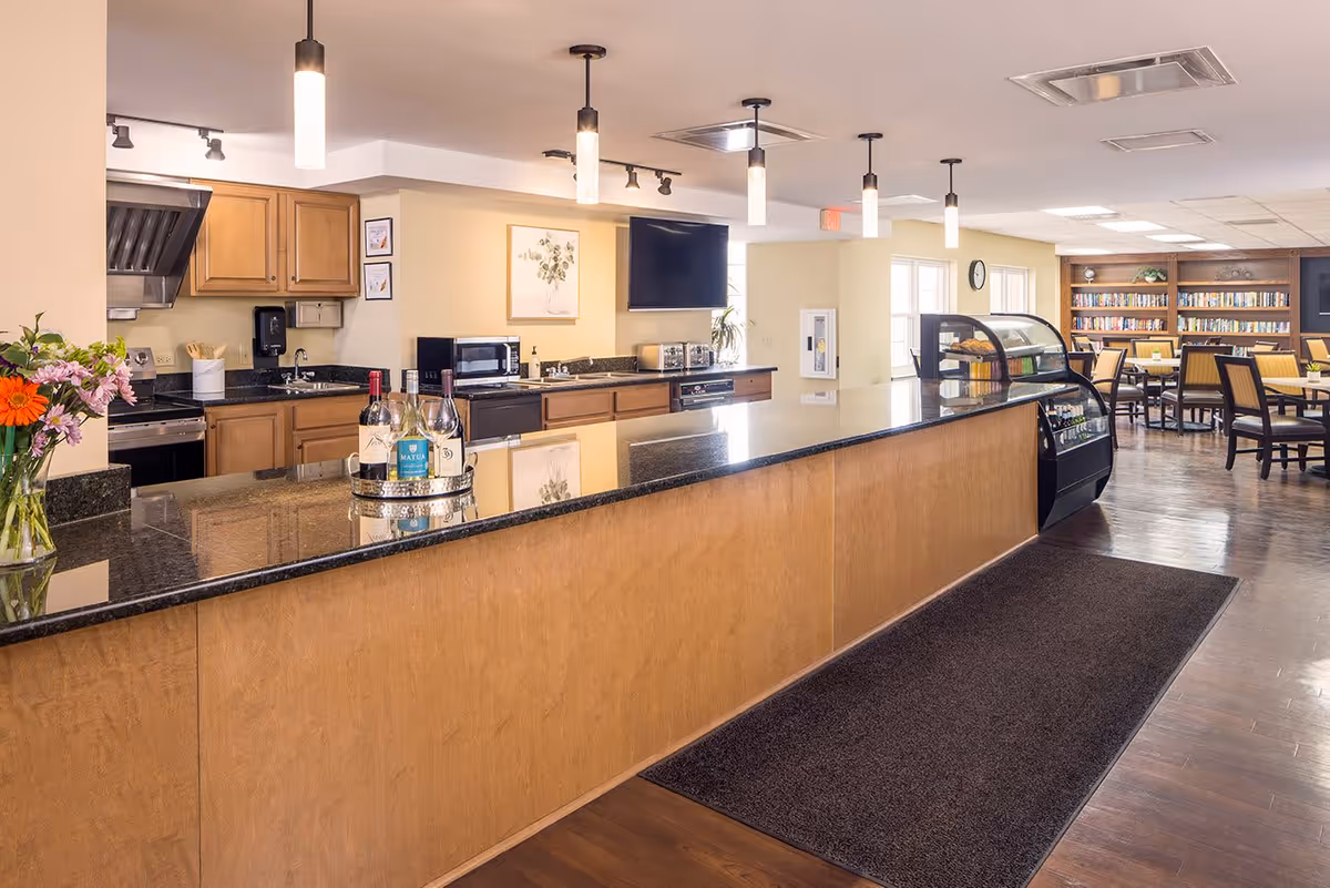 A spacious kitchen and dining area in a senior living facility with a long granite countertop, wooden cabinets, hanging pendant lights, and a display case with food items. In the background, there is a dining area with tables, chairs, bookshelves, and a television mounted on the wall.