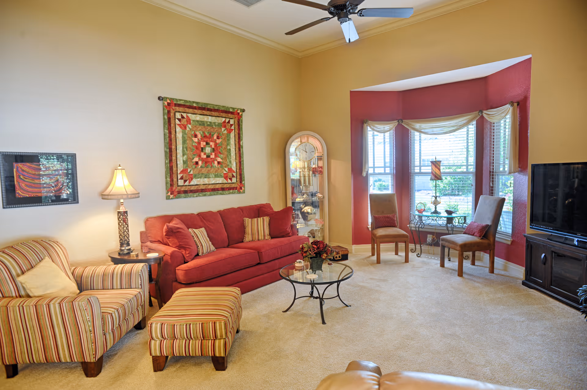 A cozy living room with a red sofa, striped armchair with matching ottoman, two wooden chairs near a bay window with blinds, a glass coffee table with a flower arrangement, a floor lamp, a wall clock in a glass case, and a flat-screen TV on a dark wooden stand. The walls are painted beige with a red accent wall around the window.