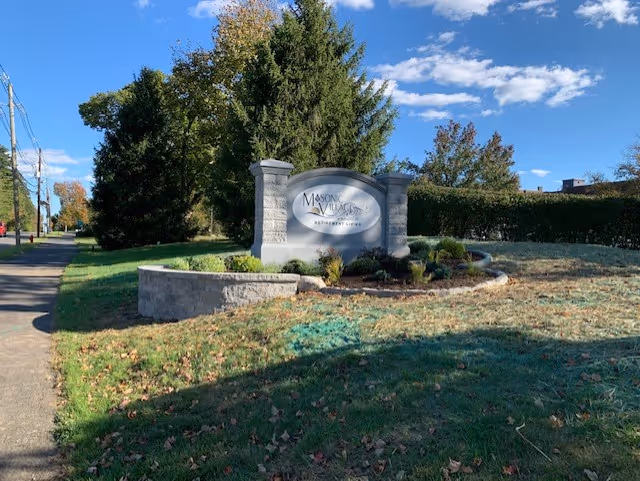Outdoor view of a stone sign for Masonic Village at Burlington surrounded by landscaped greenery and trees under a blue sky with scattered clouds.