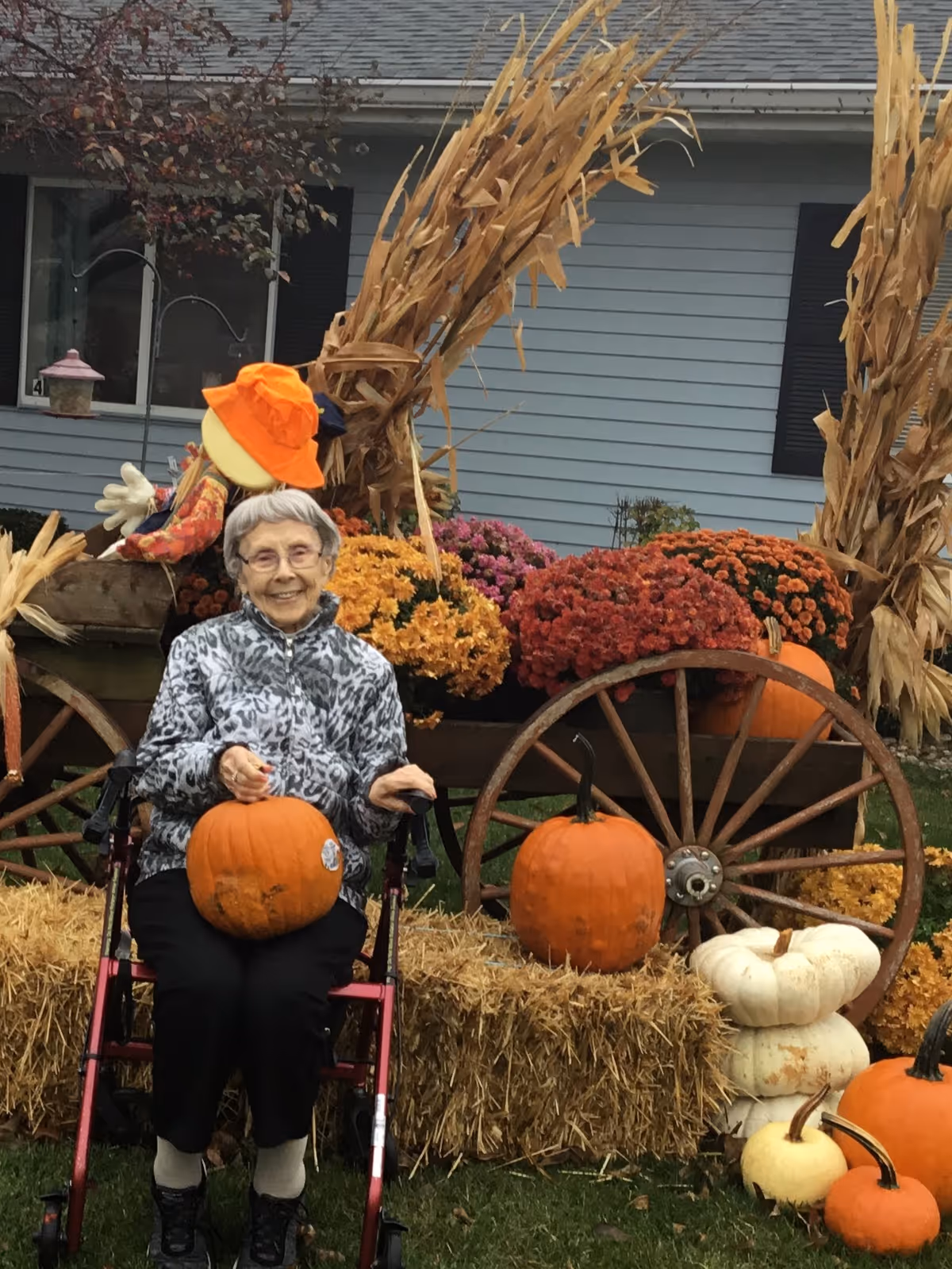 An elderly woman sitting in a wheelchair outdoors holding a pumpkin. She is surrounded by autumn decorations including hay bales, pumpkins, colorful mums, a wooden wagon wheel, and dried cornstalks. A house with blue siding and black shutters is visible in the background.