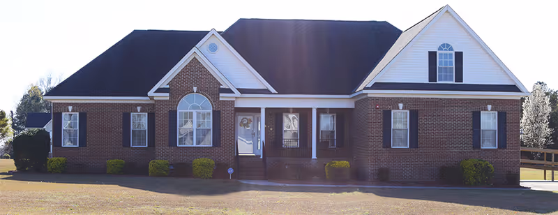 Front exterior view of a single-story brick house with a dark roof, white trim, and several windows with black shutters. There is a small porch with white columns and a front door with a wreath. The house is surrounded by a lawn with some bushes and trees in the background.